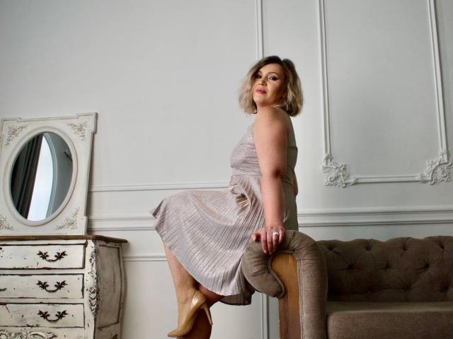 NinaStarle, met kort blond haar, draagt een mouwloze, grijze jurk en zit op een bruine, gestoffeerde bank. Ze kijkt terug met een lichte glimlach, draagt hakken en er is een witte muur op de achtergrond..