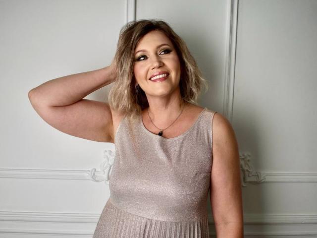 NinaStarle mit blonden, gewellten Haaren, lächelnd, trägt ein glitzerndes, ärmelloses, pinkes Kleid, eine Hand im Haar, eine kleine schwarze Halskette, mittelgroße Brüste, helle Haut, steht vor einer weißen Wand..