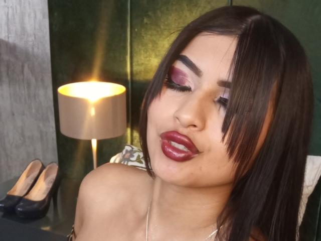 EmmaaRodriguez with dark brown hair, pink eye makeup, red lipstick, pouty lips, and a slight smile, wearing a strapless top, next to a lamp and black high heels.