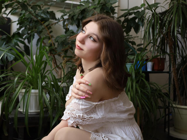 RinaLewin with wavy brown hair, looking up with a slight smile, her eyes blue, wearing a white sleeveless top, surrounded by green plants, and a gray barrier in front.