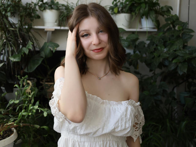 RinaLewin with medium brown hair, white off-shoulder lace top, hand in hair, slight smile, red lipstick, necklace, green-eyed, surrounded by plants. Her tits are visible.