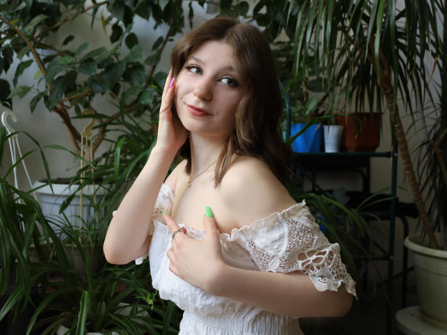 Rina Lewin, con cabello castaño claro, uñas verdes, vestido blanco de encaje con un hombro descubierto, sosteniendo sus pequeños senos, sonriendo, con las manos en las mejillas, rodeada de plantas verdes, en una habitación luminosa..