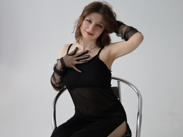 Rina Lewin mit welligem, braunem Haar, trägt ein schwarzes, durchsichtiges Kleid, schwarze Handschuhe und pinken Nagellack, berührt ihre Brüste, sitzt auf einem Metallstuhl und lächelt sanft..