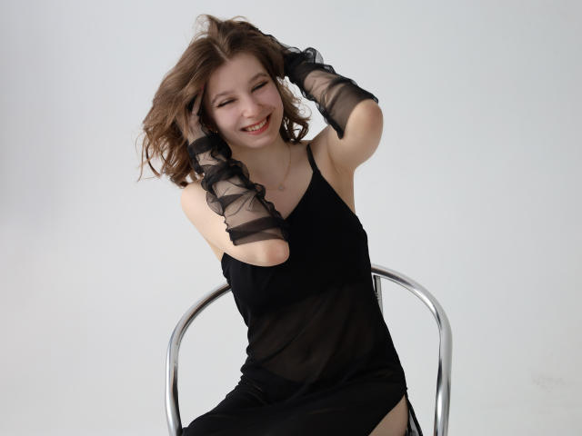 RinaLewin with wavy brown hair, smiling, wearing a sheer black dress and black mesh gloves, sitting on a metal chair, hands in hair, showing off her tits.