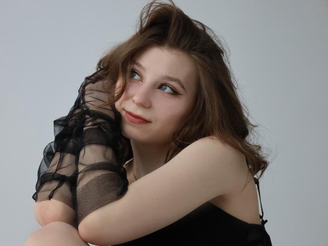 RinaLewin with wavy brown hair, blue eyes, and pale skin, smiling slightly, holding black sheer sleeves, arms crossed, wearing a black top, looking to the side.