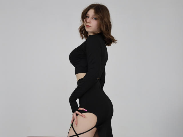 Rina Lewin, com cabelos castanho-médio, vestindo uma cropped preta de manga comprida e uma parte de baixo preta de cintura alta com recortes, que revelam suas nádegas e tiras laterais nas coxas..