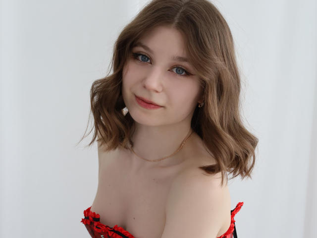 Rina Lewin, com cabelos castanhos ondulados, olhos azuis e um sutiã de renda vermelho, exibindo seus pequenos seios e pele clara, usando um colar de ouro, sorrindo suavemente..