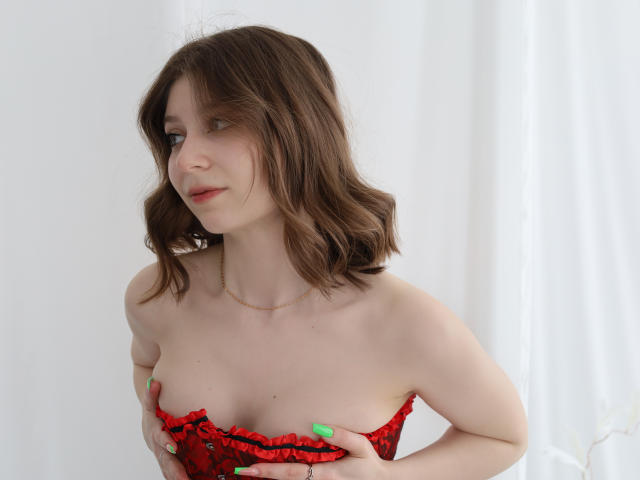 Rina Lewin, con capelli castano ondulati, una camicetta rossa arricciata, unghie verdi, che si tiene i seni, pelle chiara, un sorriso appena accennato, che guarda di lato, sfondo bianco..