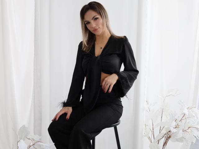 KarolinaSykes with long brown hair, black outfit, and long sleeves, sitting on a stool, hands on black pants, smiling, white background, white flowers on the right.