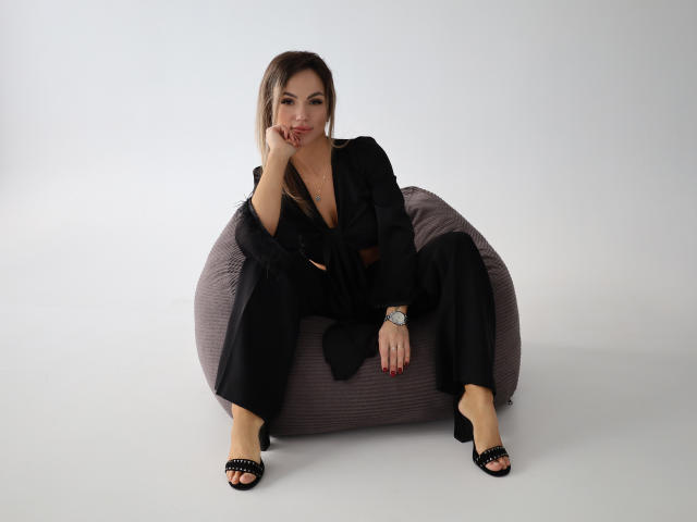 KarolinaSykes is sitting on a gray beanbag, wearing a black blazer and pants with open-toe black heels. Her brown hair is down, she's touching her chin, looking at camera.
