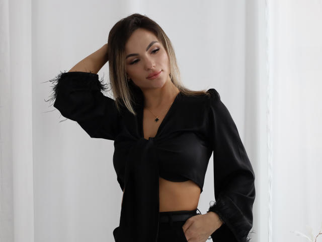 KarolinaSykes with dark hair, wearing a black crop top and matching pants, hand in hair, small tits, cute, slight smile, necklace, standing, white background.