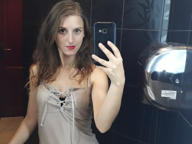 DelightBoney with wavy brown hair, red lipstick, wearing a gray lace-up top, taking a mirror selfie with a black phone in a dark-tiled bathroom. Boobs visible.