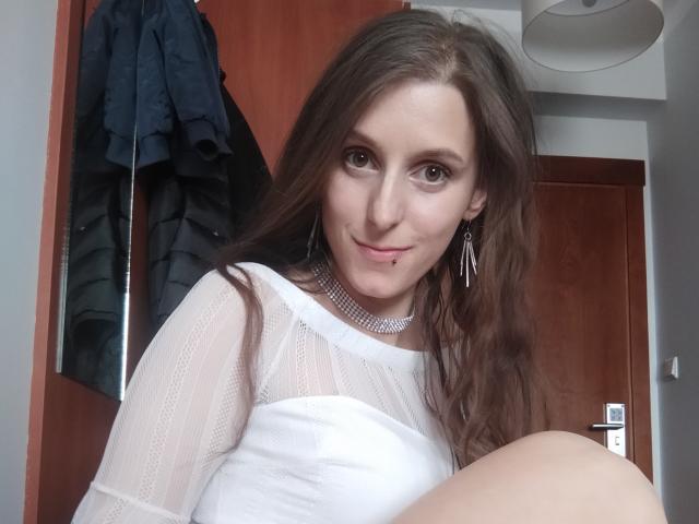 DelightBoney, with long brown hair and a white sheer top, has a nose piercing and a silver choker, smiling at the camera, showing her tits slightly.
