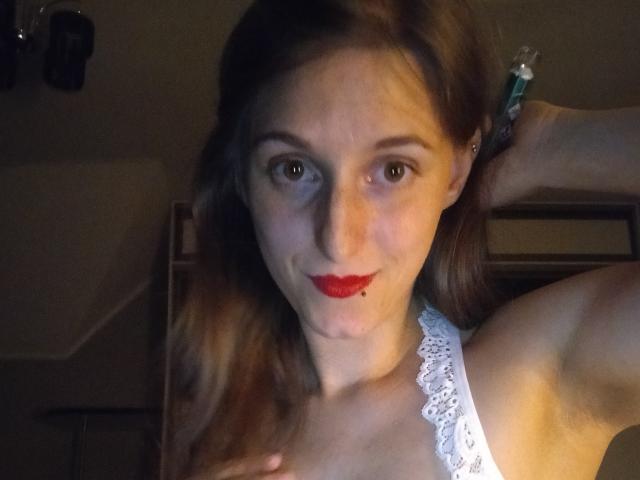 DelightBoney with red lipstick, raised arm, white lace top, brown hair, and a small bite mark on her lower lip, looking at the camera with green eyes.