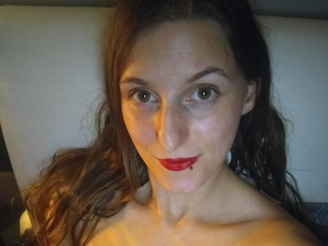 DelightBoney has red lipstick, a lip piercing, and brown wavy hair, lying down with a slight smile, her bare shoulders visible, green eyes, and a nose ring.