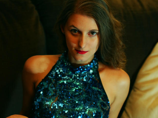 DelightBoney with wavy brown hair, red lipstick, and a blue sequined halter top, sitting on a dark couch, looking up with a slight smile. Her tits are partially visible.