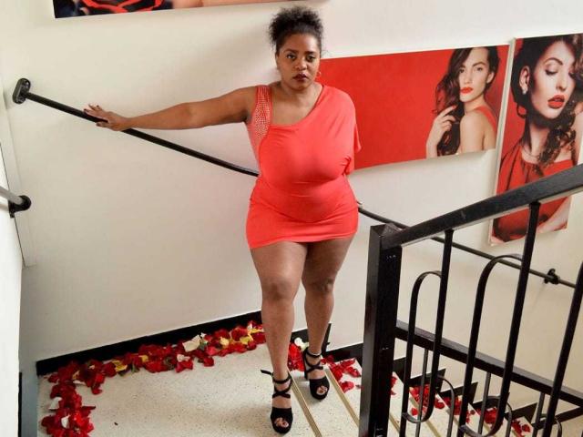 GiorgiaJhenis, curvy black woman with short curly hair, wearing a tight orange dress and black heels, standing on stairs with red rose petals, one hand on rail.