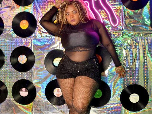 GiorgiaJhenis with curly blonde hair, black sheer top, black sparkly shorts, fishnet stockings, standing, looking at camera, neon pink background, surrounded by vinyl records.