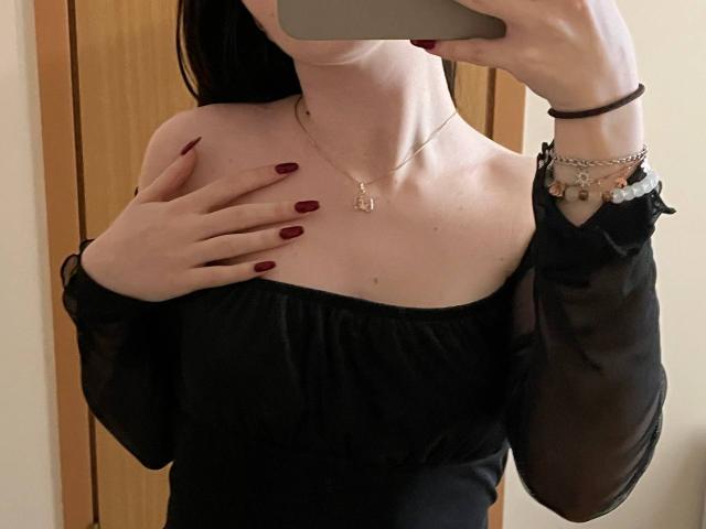 Valissia, avec une peau pâle, des ongles rouge foncé, un haut noir à une épaule, tenant un téléphone, cachant ses petites seins avec sa main, portant un collier et des bracelets, autoportrait devant un miroir..