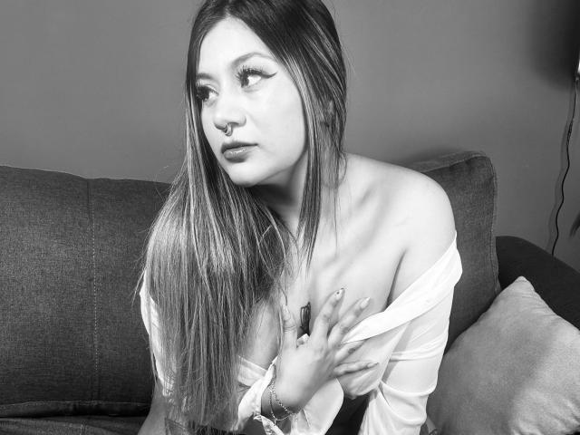 Black-and-white photo of LorraineV with long, straight hair, nose ring, and off-shoulder top, covering her small tits with one hand, sitting on a dark couch.