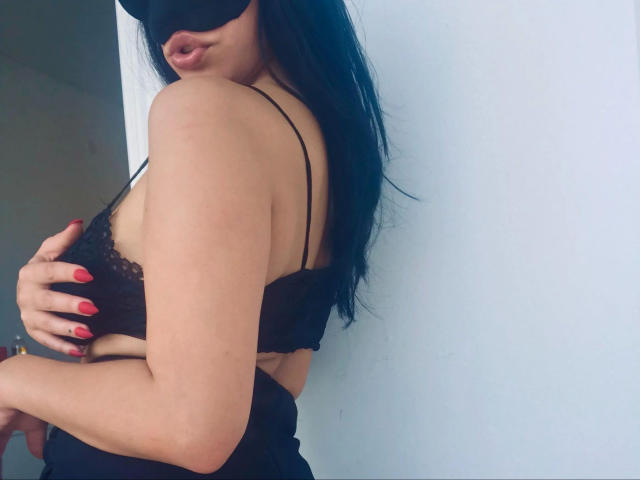 AdrianaLeone-hot with black blindfold, long black hair, red nails, black lace bra, exposing side boob, standing against white wall, pouty lips, looking over shoulder, hand on breast.