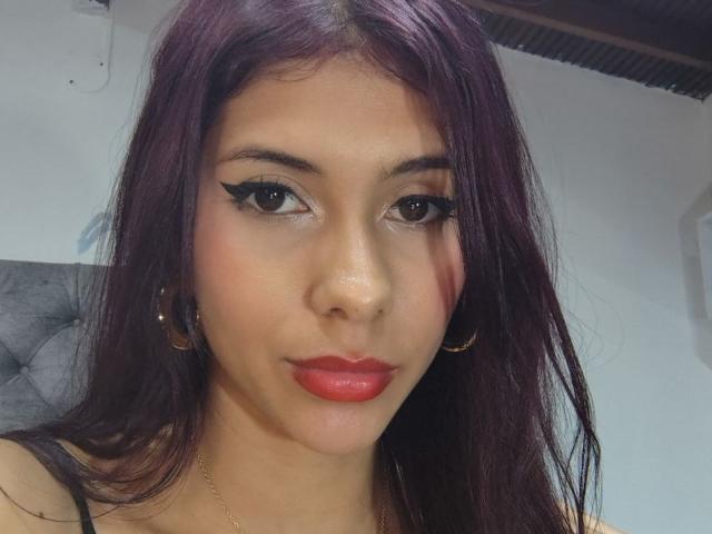 DanikaSerov has dark purple hair, brown eyes with thick eyeliner, red lipstick, and gold hoop earrings. Her skin is light brown, and she's wearing a gold necklace.