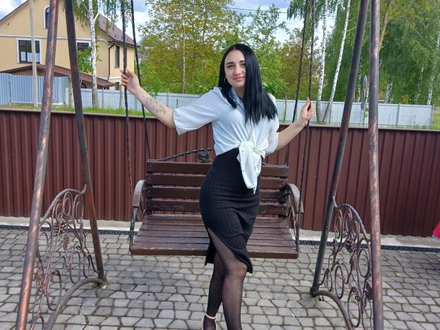 DeliaLiv with black hair, white blouse tied at the waist, black skirt, and black stockings, standing on a wooden swing, arms out, tattoo on left arm, cobblestone ground.