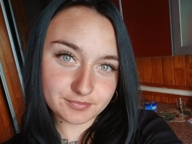 DeliaLiv has black hair, green eyes, and a small smile. She's wearing a black shirt and gold hoop earrings. Her skin is fair with a slight tan.