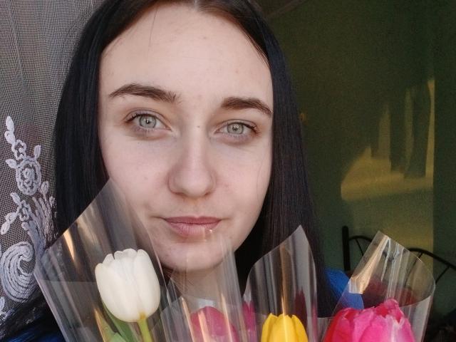 DeliaLiv with pale skin, green eyes, and dark hair, holding a bouquet of white, yellow, pink, and red flowers, smiling subtly. She's in front of a semi-transparent window.