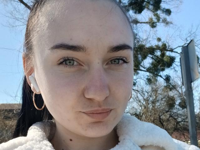 DeliaLiv has pale skin, green eyes, and dark eyebrows. She's wearing a white fluffy jacket, gold hoop earrings, and white earbuds. Her black hair is tied back.