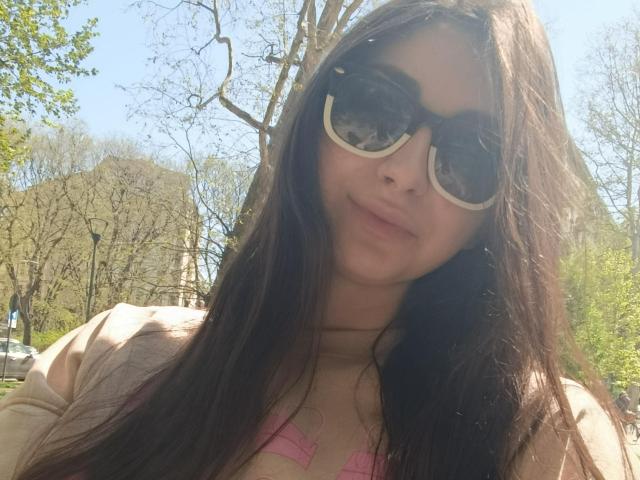 CandiceJaguar with long brown hair, wearing big black sunglasses, and a pink bra, has a subtle smile, and is standing in a sunny, tree-filled area.