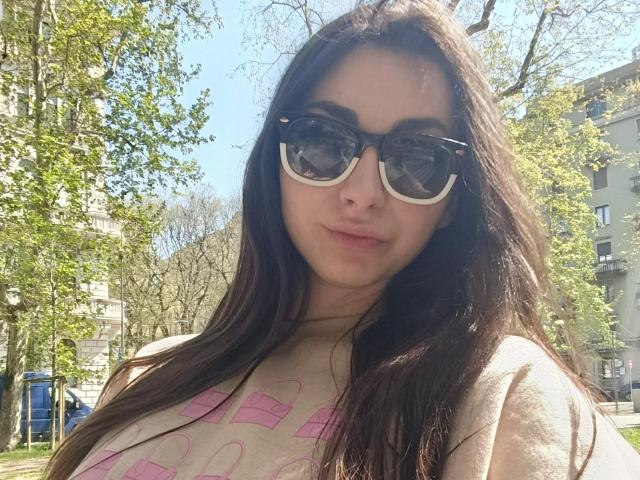 CandiceJaguar with long dark brown hair, wearing black and white sunglasses, and a beige shirt with pink patterns, standing outside in sunlight. Boobs slightly visible under shirt.