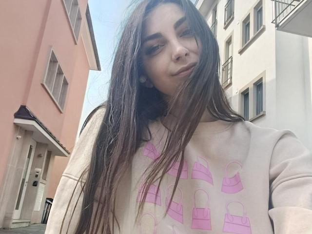 CandiceJaguar with long brown hair, wearing a pink purse-print sweater, has a subtle smile, and is standing in a narrow urban alleyway with pink and white buildings.