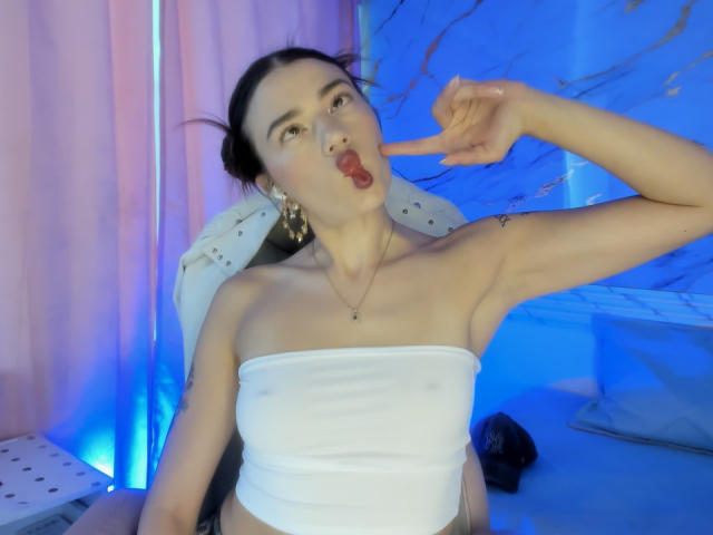 JasmineHypery with dark hair in a bun, white tube top, red lipstick, and gold earrings, sticking her tongue out, pointing at her mouth, showing a tattoo on her arm.