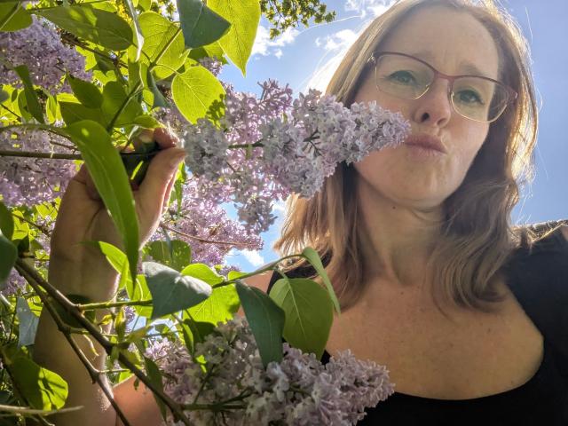 HeisseLina-hot with glasses, pouty face, and light brown hair, holding lilac flowers, her black top showing her tits, green leaves, and sunlight.