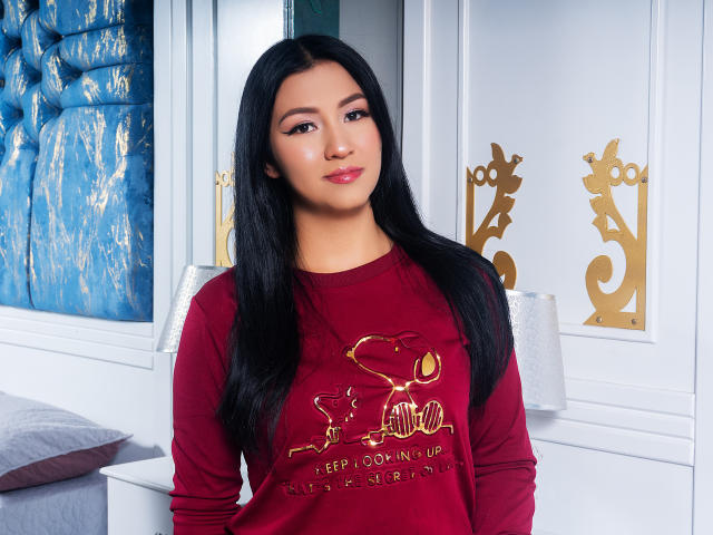 JulianaWonder with long black hair, red sweatshirt, and gold heart design, standing in a room with white and blue decor. Her ass is not visible. She has a cute smile.