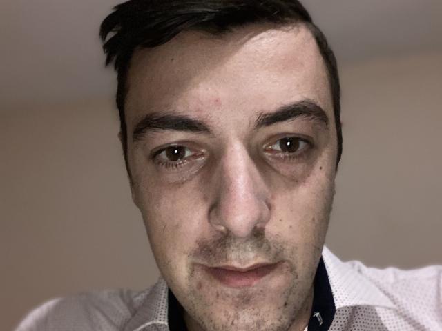 MaxenRace has a slightly puffy face with brown eyes, dark eyebrows, and short dark hair. He's wearing a white shirt with a black collar. His skin is pale.