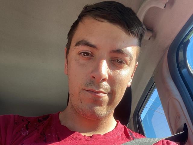 MaxenRace has a slight tan, short brown hair, and is wearing a red shirt with a black seatbelt across his chest, sitting in a sunny car.