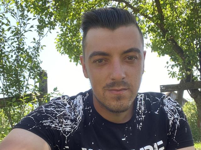 MaxenRace, a dude with short, slicked-back black hair, stubble, and brown eyes, wearing a black graphic tee, looking at the camera with a slight smile.