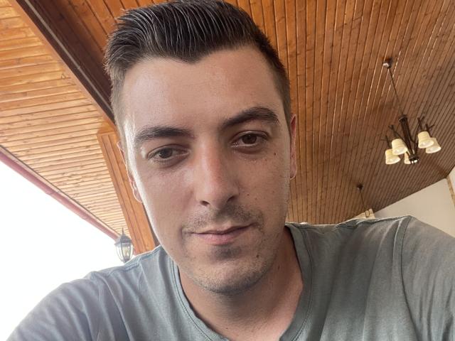 MaxenRace has short, dark, slicked-back hair, a light green V-neck shirt, and a small earring in his left ear, looking slightly down with a neutral expression.