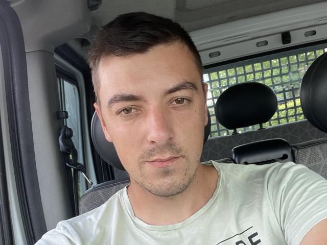 MaxenRace is a dude with short brown hair, light green eyes, and a slight beard, wearing a white tee, sitting in a car, looking at the camera.