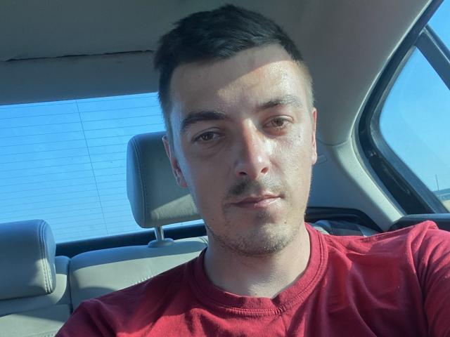 MaxenRace has a red shirt, short dark hair, and a stubble. He's sitting in a car with grey headrests, looking at the camera with a neutral expression.