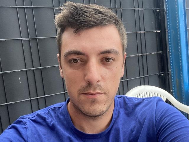 MaxenRace, a medium-build dude with short brown hair, wearing a blue shirt, has a serious expression, brown eyes, and a light beard. He's sitting on a white chair.