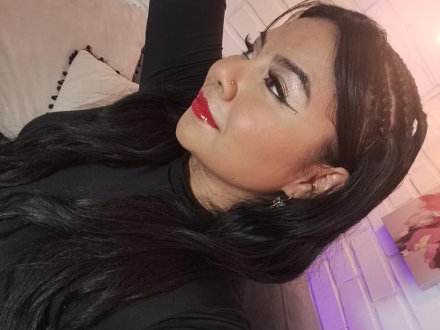 ValentinaSilva with shiny black hair, red lipstick, and dramatic eyeliner, wearing a black top, nose piercing, and star earrings, looking to the side with a slight smile.