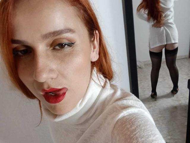 SelenneMillers with red hair, white turtleneck, red lipstick, and black thigh-highs, showing her ass in a mirror reflection, wearing black heels. Her face is in the foreground.
