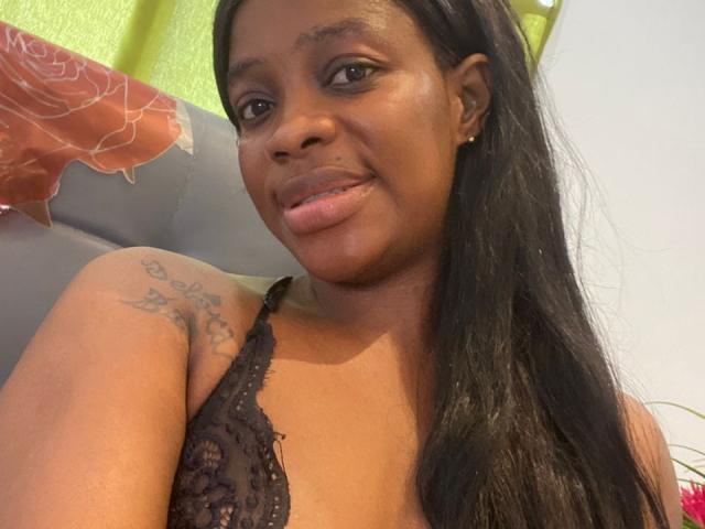 MELLEE tem a pele de cor marrom escura, cabelos pretos longos e uma tatuagem no ombro direito. Ela está vestindo uma blusa preta de renda, sorrindo com os lábios ligeiramente entreabertos..
