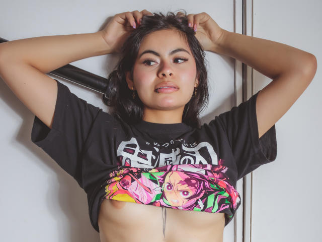 CrystallThonpsom with hands in black hair, wearing black t-shirt, pink and green graphic, tits exposed, arms up, pink nails, neutral expression, white background, medium shot, photo.