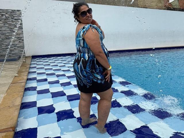 Keyla69Hot, chubby with dark curly hair, wearing a blue patterned dress, black sunglasses, standing in a blue and white checkered pool, water splashing, looking back, wet skin.