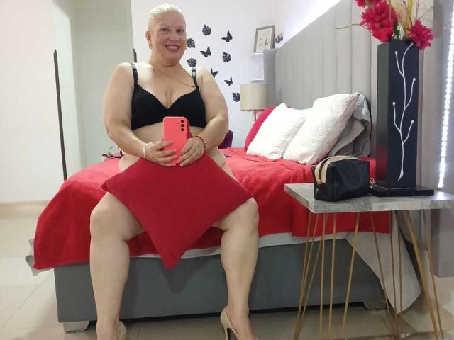 RebeccaSinss, formosa con la pelle chiara, capelli biondi corti, reggiseno nero, che tiene un cuscino rosso, seduta sul letto, coperta rossa, tacchi beige, telefono rosso, sorridente, camera da letto, comodino con borsa nera..