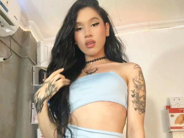 EnchantressKanne with long black hair, wearing a light blue strapless top, showing tattoos on her arm and chest, and a black choker, with her tongue slightly out.